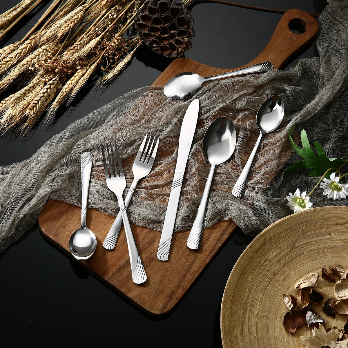 A wooden board displays a 48-piece stainless steel flatware set with various spoons and forks on a gray cloth.
