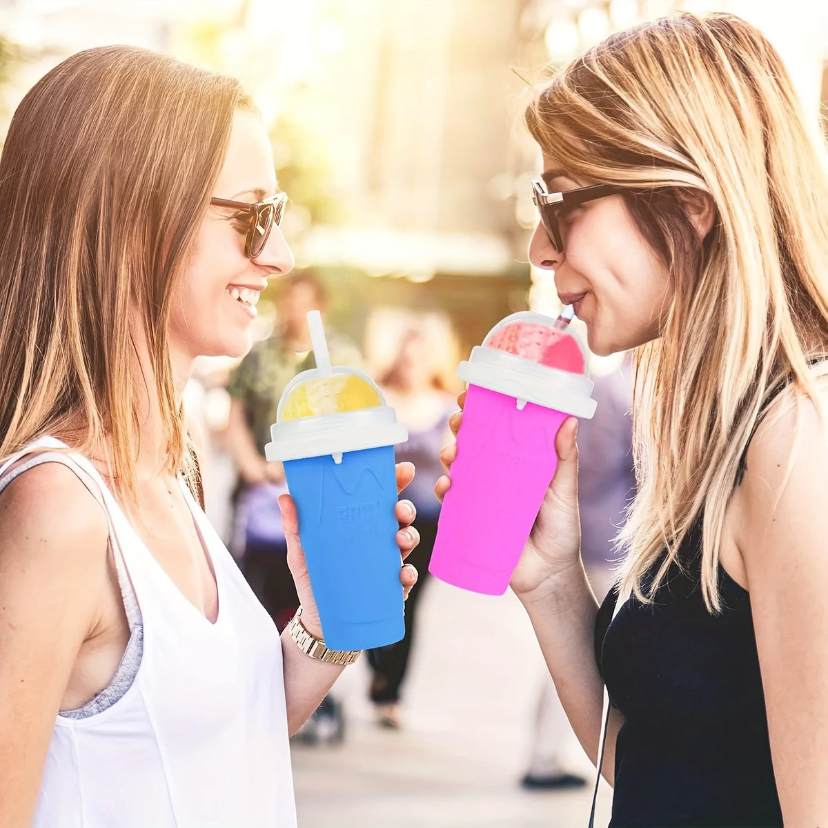 Two women smiling while holding and drinking colorful DIY slushy maker cups outdoors