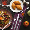 A flat lay showing a 48-piece stainless steel flatware set with croissants and fruit on a dark background.