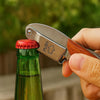 A hand holds a silver wine opener with a wooden handle and leather case, positioned near a green glass bottle.