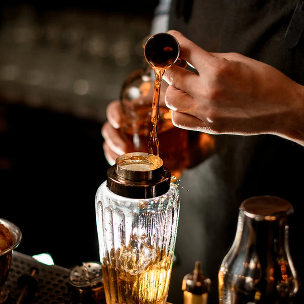 Eine Hand gießt bernsteinfarbene Flüssigkeit aus einem Jigger in ein Glas vor dunklem Hintergrund.
