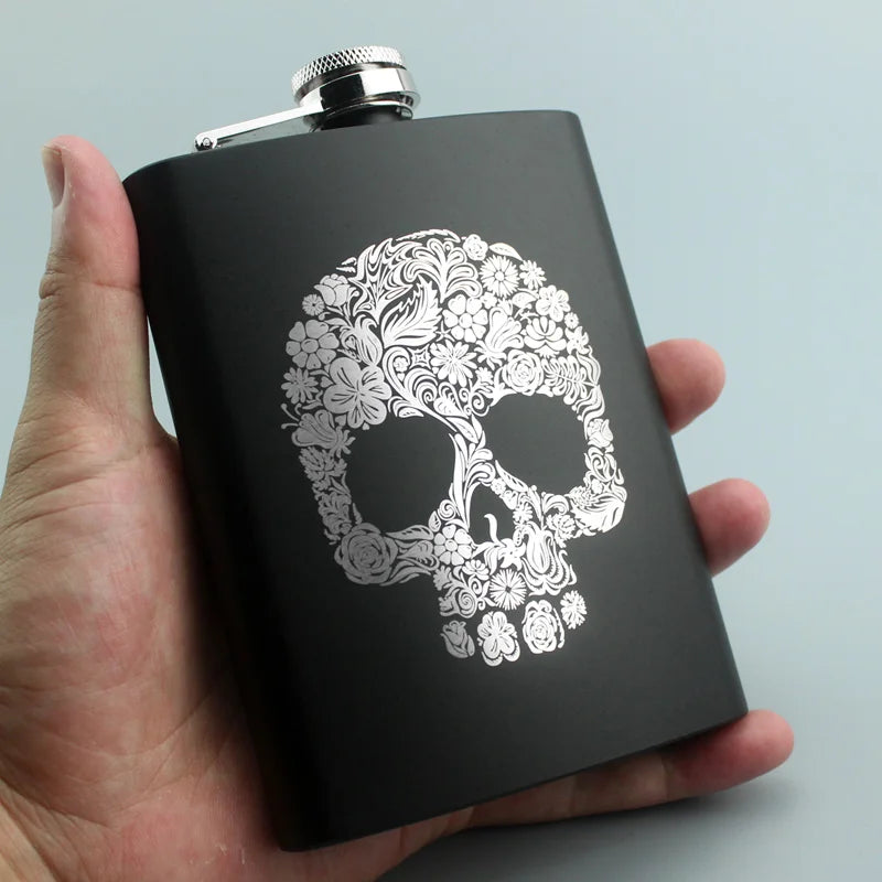 A hand holds a black laser engraved stainless steel hip flask with a decorative skull design.