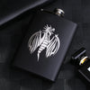 A black laser engraved stainless steel hip flask with a silver cap resting on a dark textured surface.