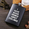A black laser engraved stainless steel hip flask resting on a wooden surface next to a golden Eiffel Tower and a box.