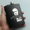 A hand holds a black laser engraved stainless steel hip flask with a silver cap.