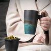 Una persona sostiene una taza de té de cerámica oscura con tapa, junto a una chaqueta beige claro y un libro.