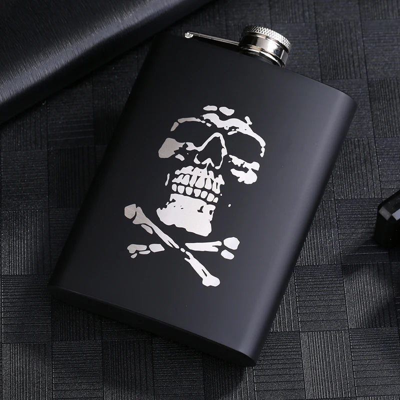 A black laser engraved stainless steel hip flask with a silver cap resting on a dark textured surface.
