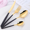 A set of elegant gold and black stainless steel flatware including a knife, fork, spoon, and soup spoon on a white plate.