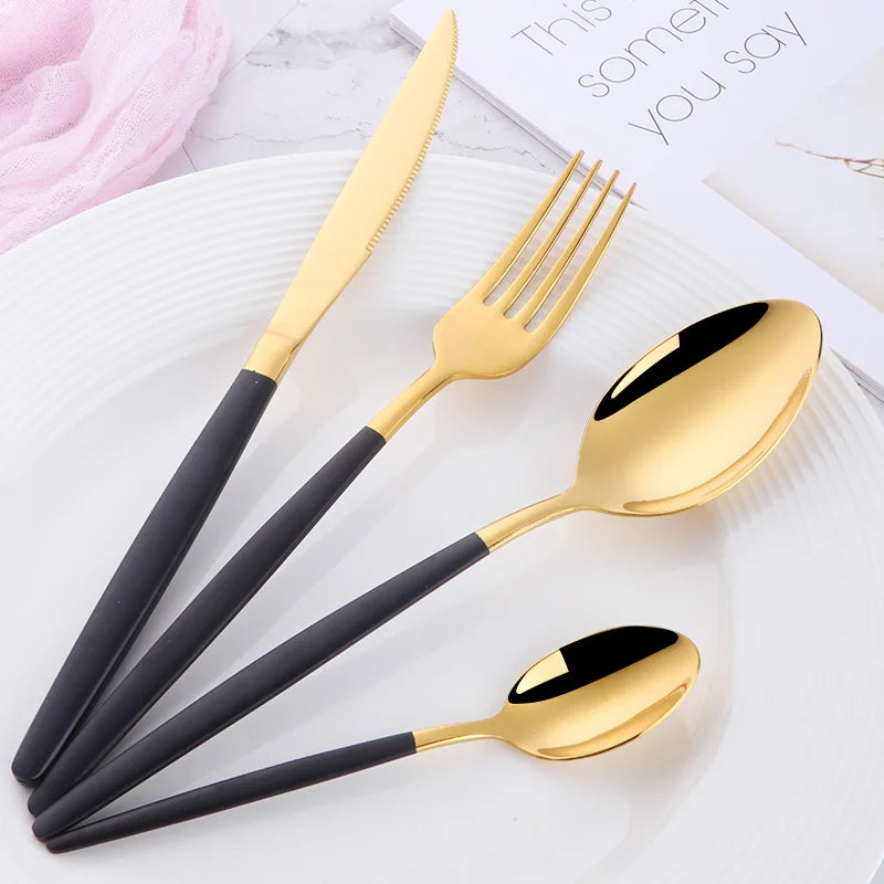 A set of elegant gold and black stainless steel flatware including a knife, fork, spoon, and soup spoon on a white plate.