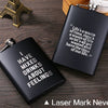 A black laser marked stainless steel hip flask with text about feelings resting on a wooden surface.