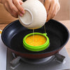 Hands are pouring liquid from a white silicone ring into an orange-yellow mixture in a black frying pan.