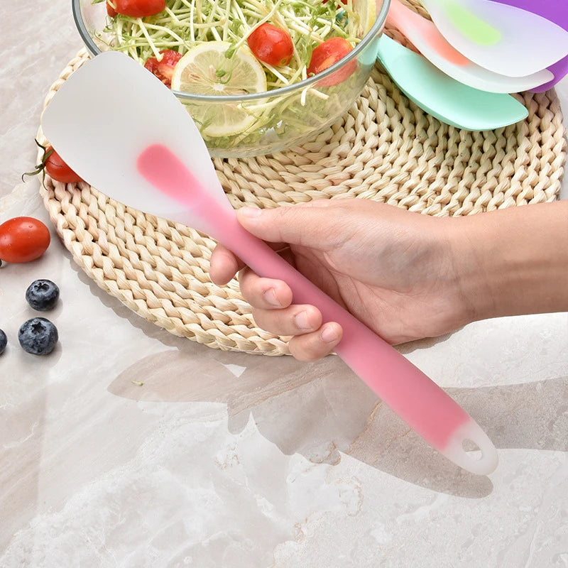 A hand holds a pink silicone cooking spatula over a bowl of salad and a set of colorful silicone spatulas.