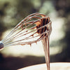 A close-up of a stainless steel balloon whisk showing its wire loops against a blurred outdoor background.