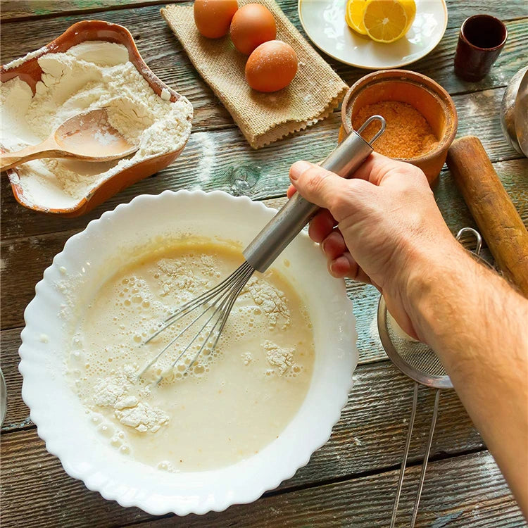A hand uses a stainless steel balloon whisk to mix liquid in a white bowl, with eggs and spices nearby.