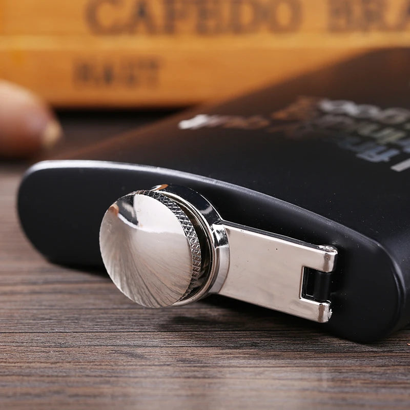 A close-up of a black hip flask with a silver laser engraved stainless steel cap resting on a wooden surface.