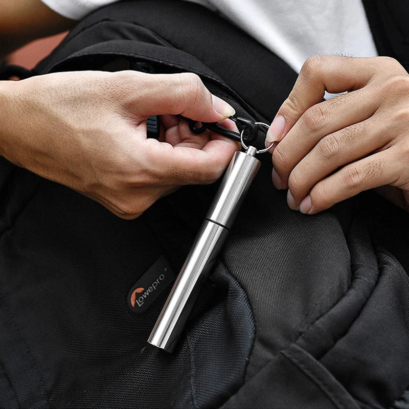 A hand is holding a stainless steel telescopic drinking straw with a cleaning brush against a black fabric background.