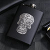A black laser engraved stainless steel hip flask with an intricate skull design resting on a dark textured surface.