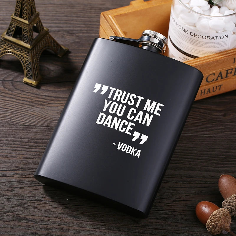 A black laser engraved stainless steel hip flask with a silver cap resting on a wooden surface next to decorative items.