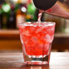 A close-up shot of a cocktail being poured into a glass filled with ice and red liquid, held by a stainless steel shaker.