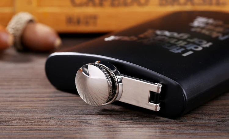 A black hip flask with a silver-colored engraved closure is resting on a wooden surface.