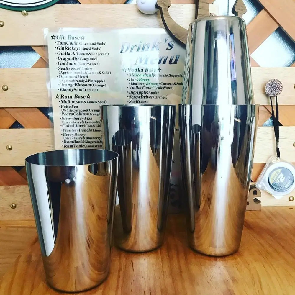 A set of professional stainless steel cocktail shakers and various drink ingredients displayed on a wooden surface.