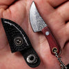 A hand holds a set of Damascus pattern pocket mini knives, featuring black handles and silver accents.