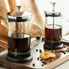 A glass and stainless steel coffee press pot filled with dark brewed coffee sits on a wooden tray.