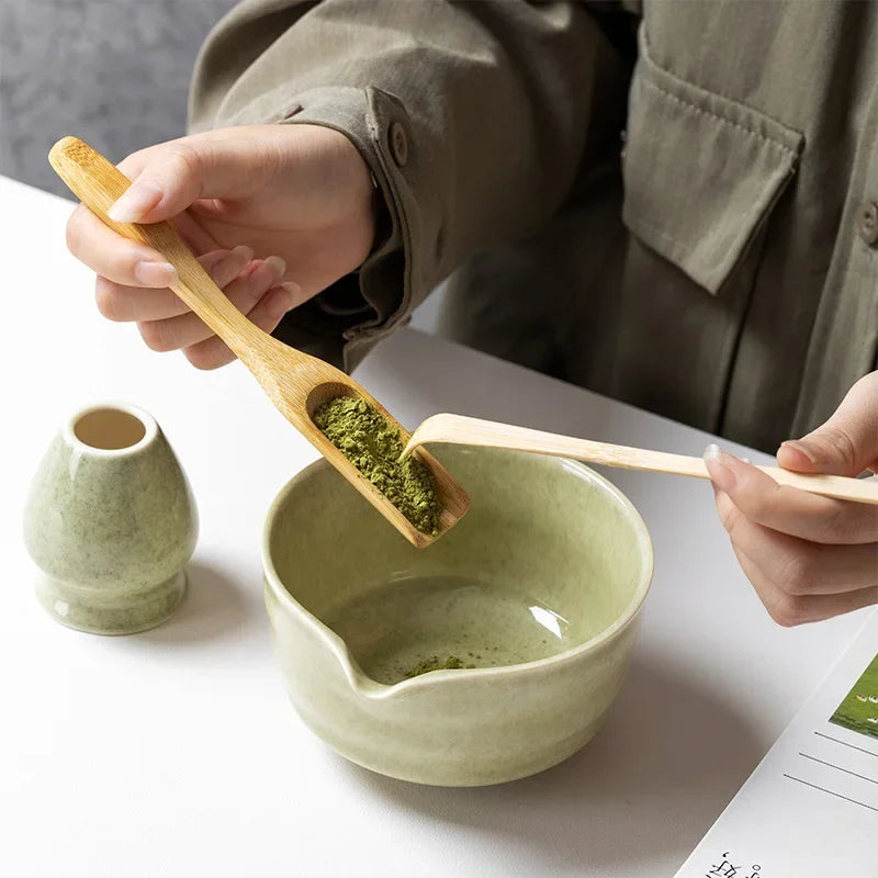 Una mano usa una cuchara de madera para recoger polvo de matcha verde en un tazón de cerámica verde claro.
