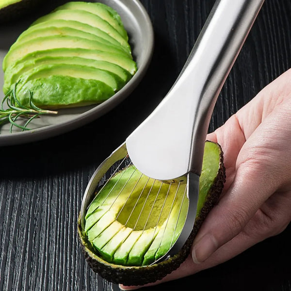 A hand holds a stainless steel avocado cutter slicing an avocado, showing the green flesh and pit.