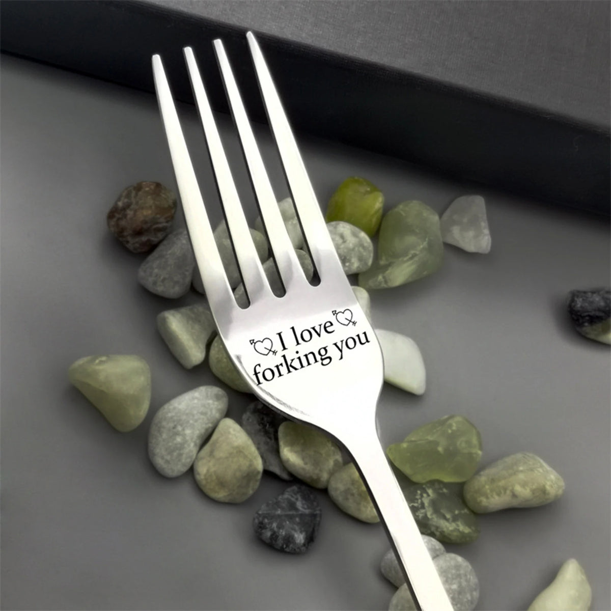 A silver stainless steel fork engraved with the text I love forking you resting on a bed of smooth green and gray stones.