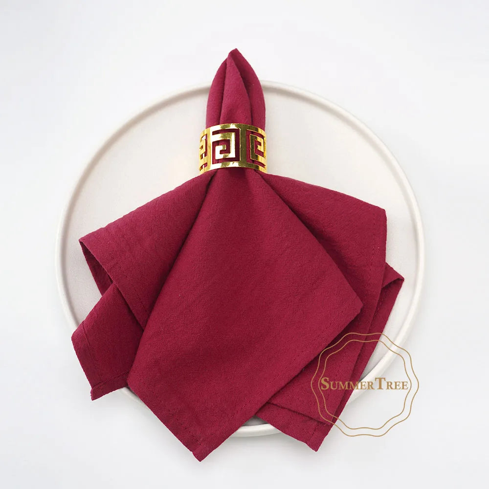 A stack of ten deep red cotton table napkins tied with a gold geometric ribbon, displayed on a white plate.
