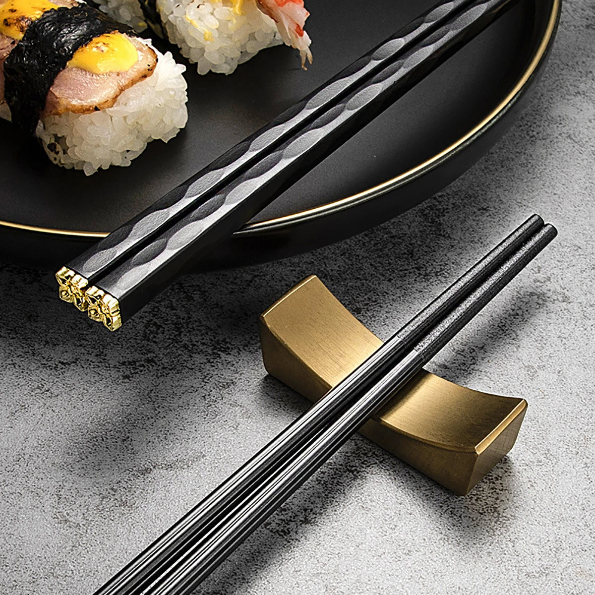 A set of five black and gold alloy chopsticks resting on a dark plate next to sushi rolls.