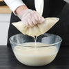 A hand wearing a glove is dripping liquid from a mesh strainer into a glass bowl.