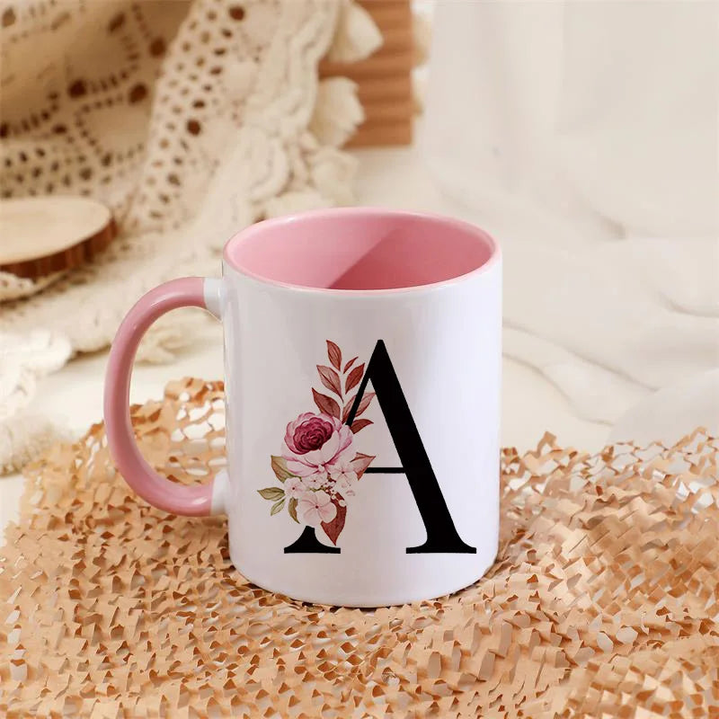 A white coffee mug with a pink handle featuring a large black letter A decorated with pink and green floral elements.