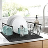 A black silicone mat holds various kitchen utensils and cups on a countertop near a window.