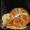 A stainless steel and wood dough whisk set resting on a wooden board with baked bread.