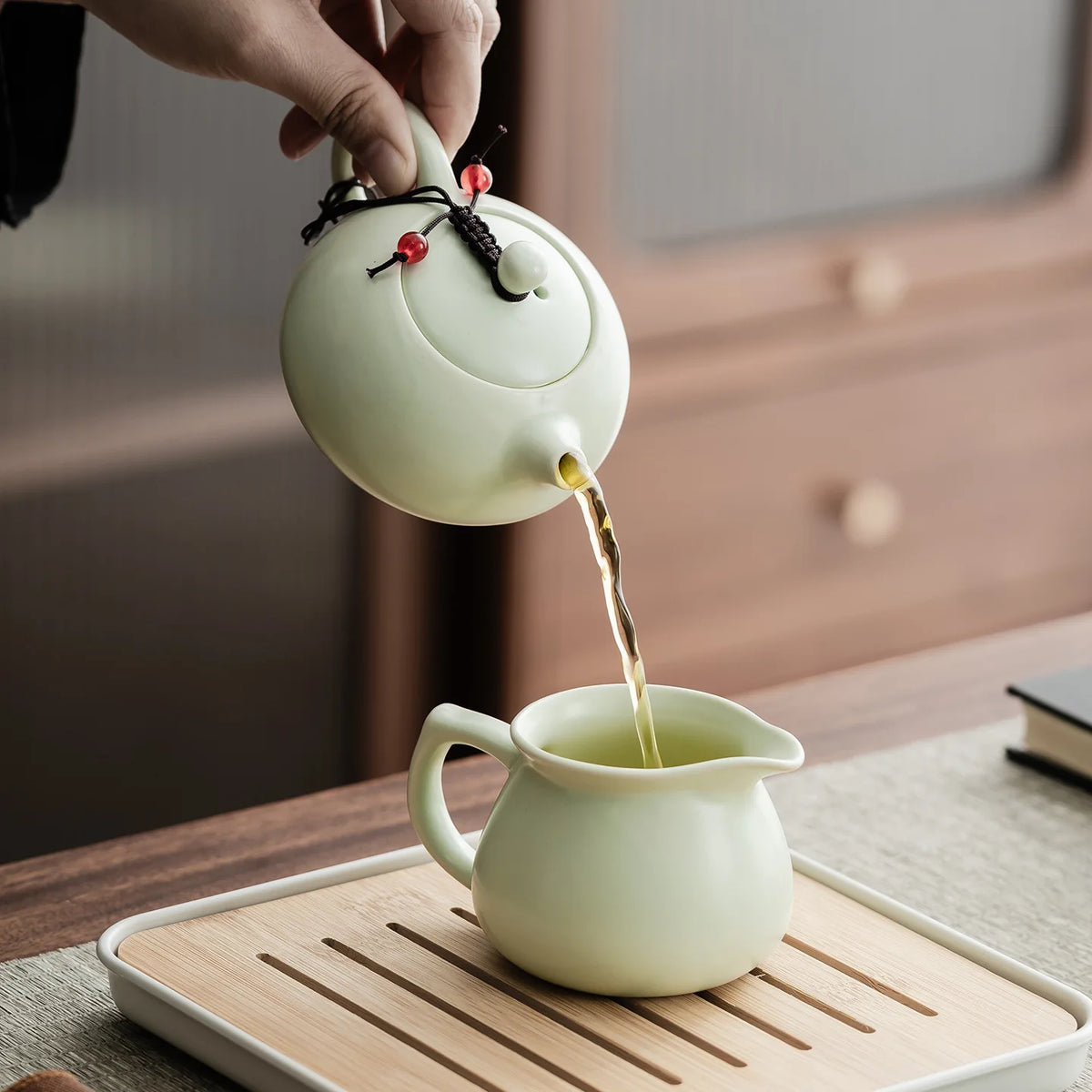 A hand pours tea from an elegant ceramic teapot into a cup on a wooden tray.