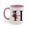 A pink coffee mug with a large black initial H decorated with purple roses and green leaves.