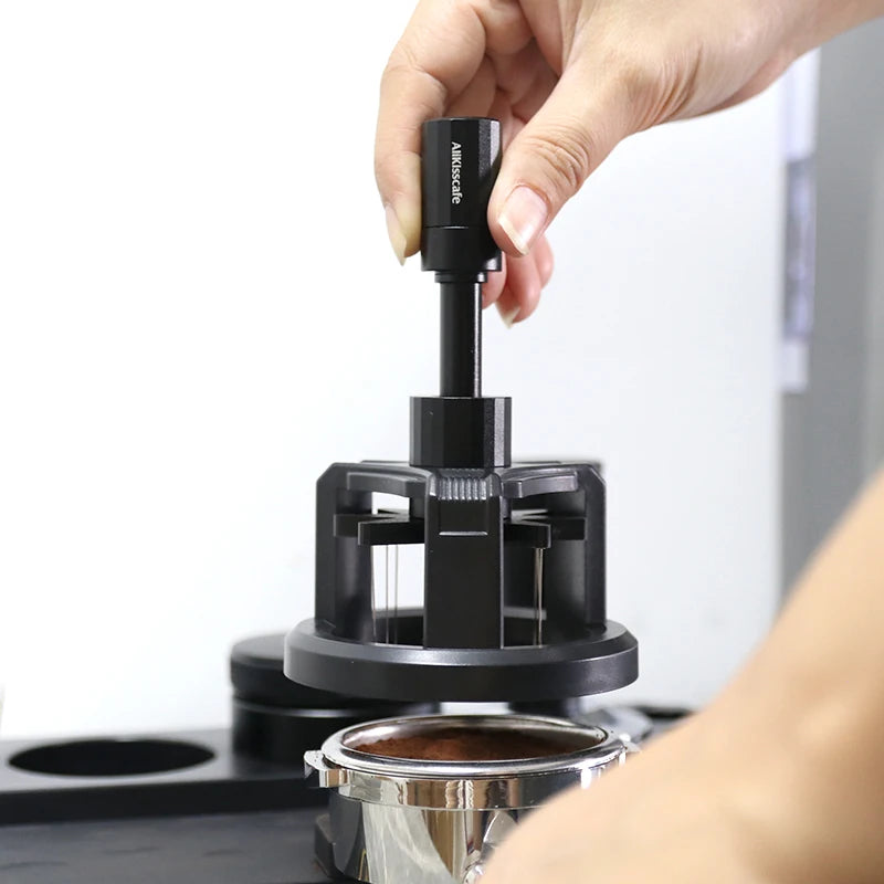 Une main tient un outil WDT noir professionnel pour remuer l'espresso, positionné au-dessus d'un récipient métallique.