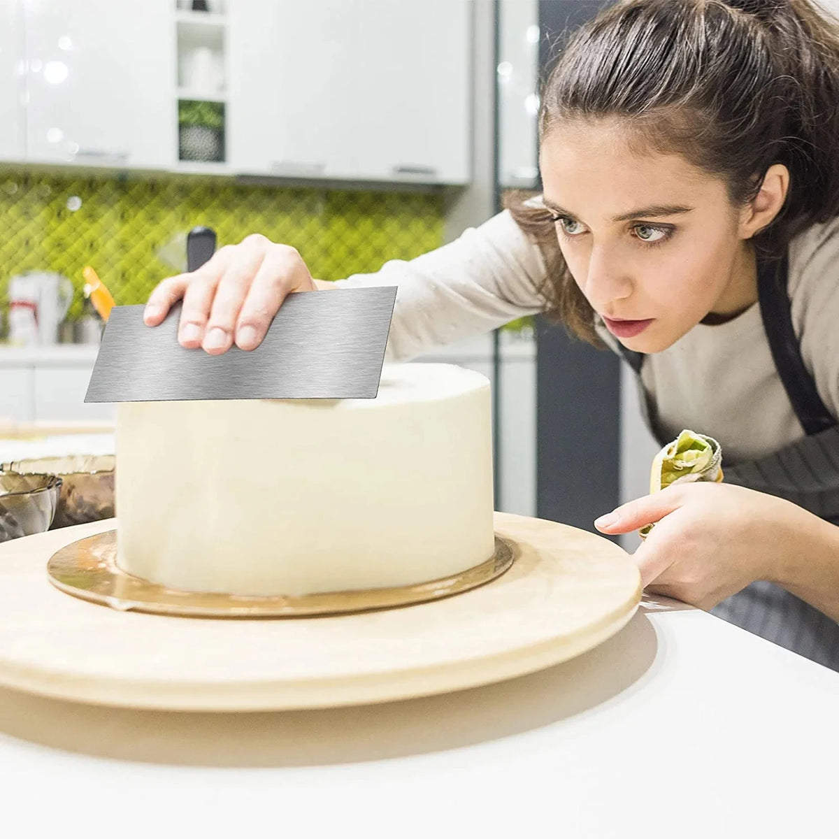 A woman uses a stainless steel cake decorating scraper to smooth the side of a white cake in a kitchen setting.