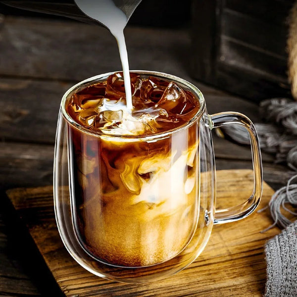 Nahaufnahme einer doppelwandigen isolierten Glas-Kaffeetasse, die mit Milch gefüllt wird und auf einer Holzoberfläche ruht.