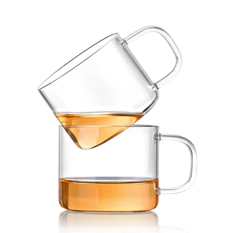 A clear glass tea cup set showing a pour of amber tea in the upper cup and liquid in the lower mug.