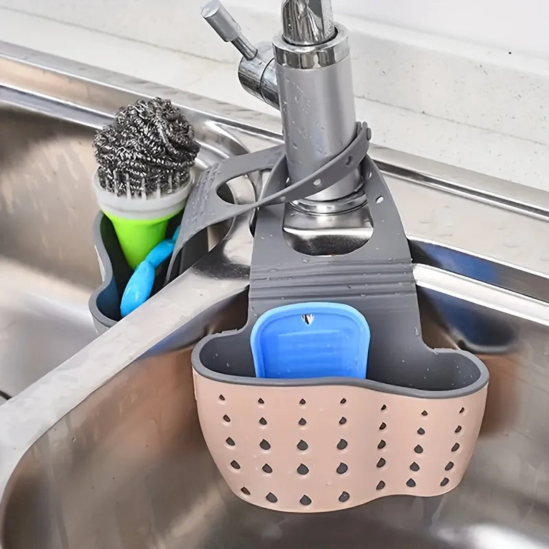 A gray hanging sponge rack with a blue holder and a green brush is placed in a stainless steel kitchen sink.