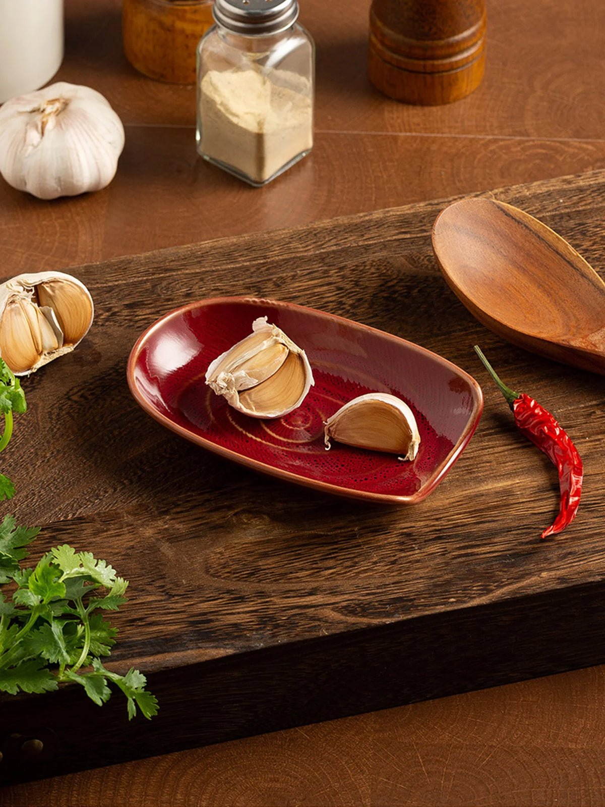 A wooden cutting board displays garlic cloves, a red chili pepper, and a glass jar of spice on a dark surface.
