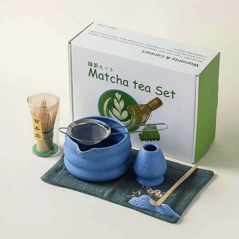 A box containing a traditional Japanese matcha tea set with a bamboo whisk, strainer, and bowl on a green cloth.