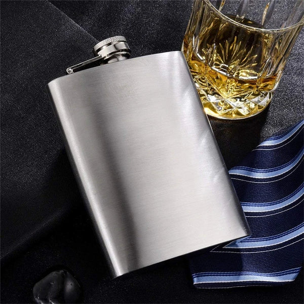 Silberne Edelstahlflasche mit Metalltrichter auf dunkler Oberfläche neben einem Glas bernsteinfarbiges Getränk.