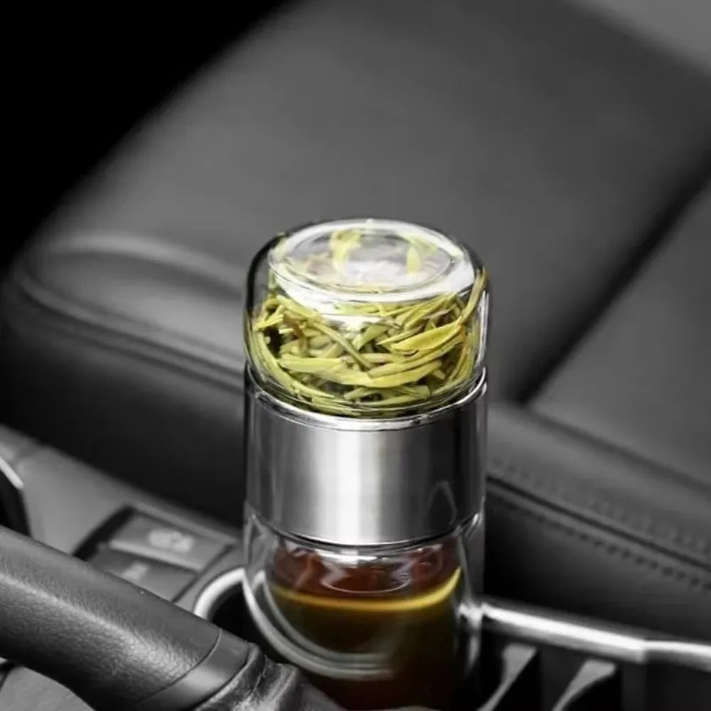 A glass tea infuser filled with dried green tea leaves sits on a dark surface.