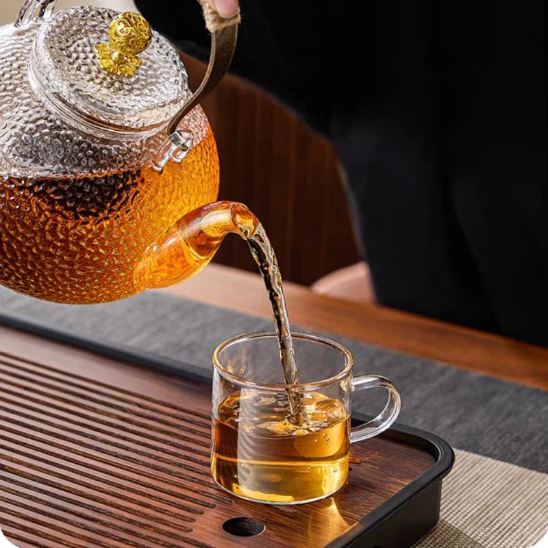 A hand-hammered glass teapot pours tea into a glass cup on a wooden tray.