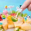 A hand holds a white cat-shaped fruit and dessert pick surrounded by various colorful fruit and sweet treats.