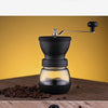 A manual coffee grinder with a glass burr and sealed storage container is shown on a wooden surface.